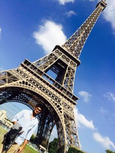 Men standing Infront of an Eiffel Tower holding a jacket in his hands under bright blue sky.